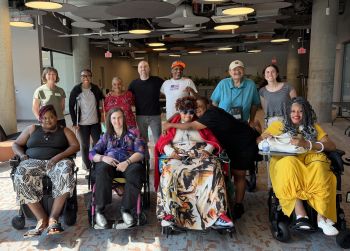 A group of twelve people facing the camera and smiling. The four people in the front chairs are in wheelchairs, one member of the backrow has a walker, and two others in the backrow have white canes.