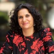 A headshot of Dr. Sarah Dababnah. She has dark curly hair and is smiling at the camera. She is wearing a black and red top.