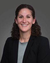 A picture of Dr. Julie Rekant. She is a white woman with dark curly hair. She is smiling facing the camera wearing a black blazer over a light blue shirt.