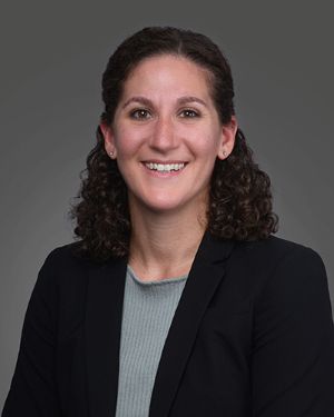 A picture of Dr. Julie Rekant. She is a white woman with dark curly hair. She is smiling facing the camera wearing a black blazer over a light blue shirt.