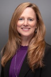 A white woman with long blond hair and blue eyes is smiling at the camera. She is wearing a purple top.